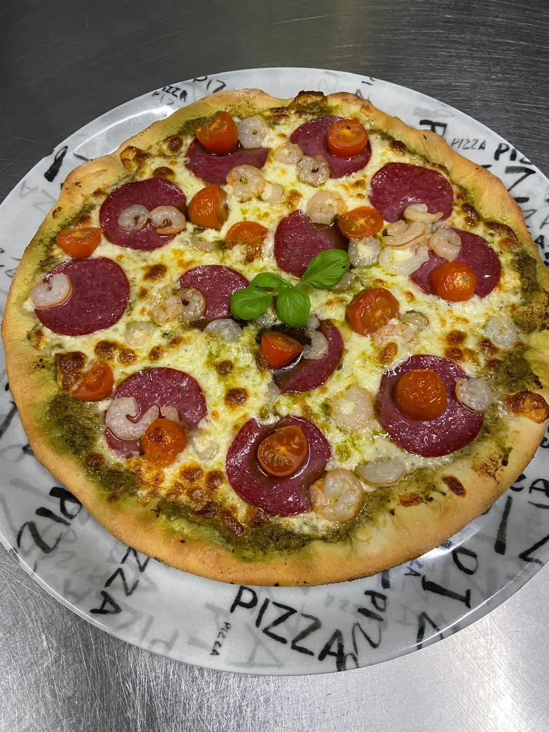 Pizza mit Salami, Garnelen, Cocktailtomaten und Basilikum auf dem Pizzateller des La Mamma Mia